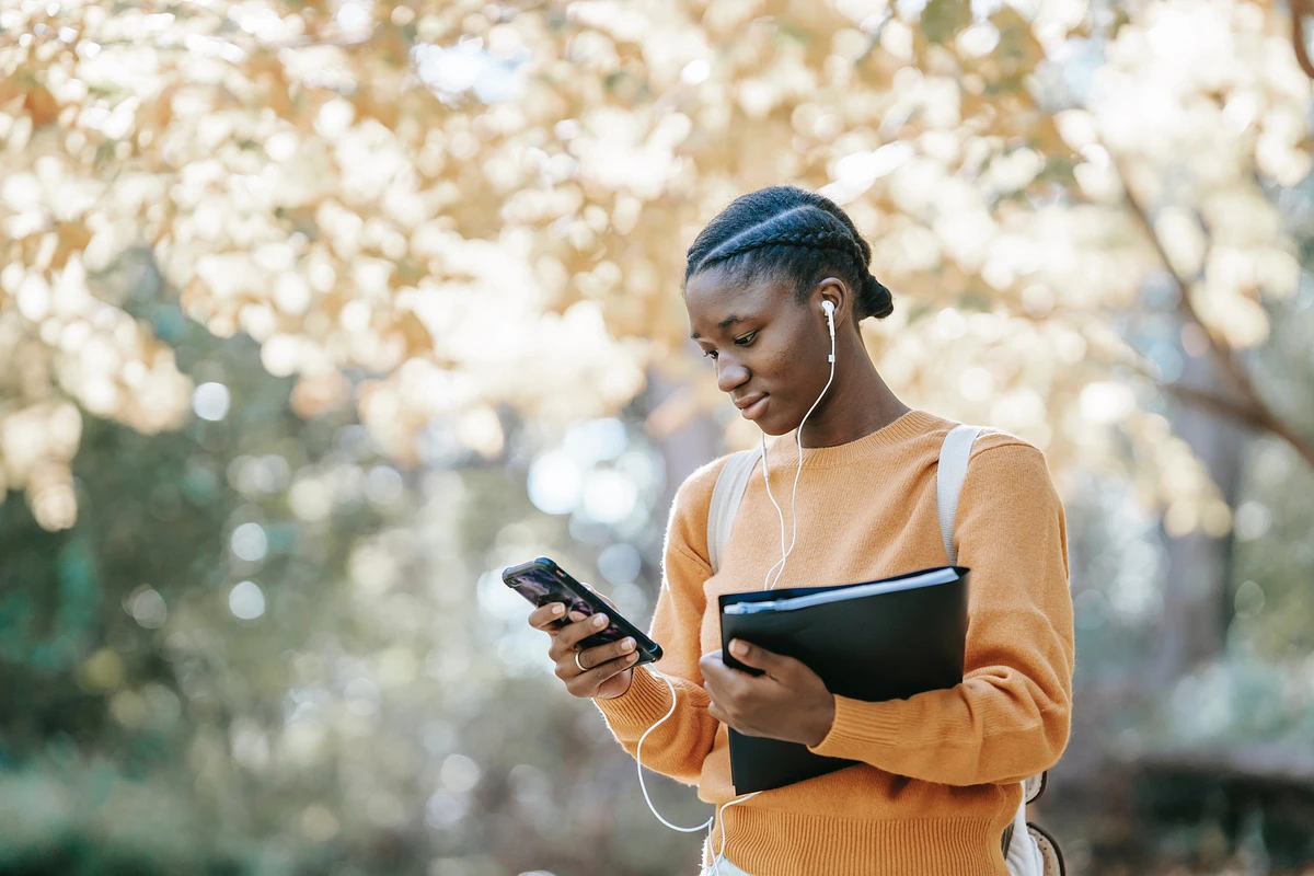 A student walks under a tree carrying a book and an iPhone